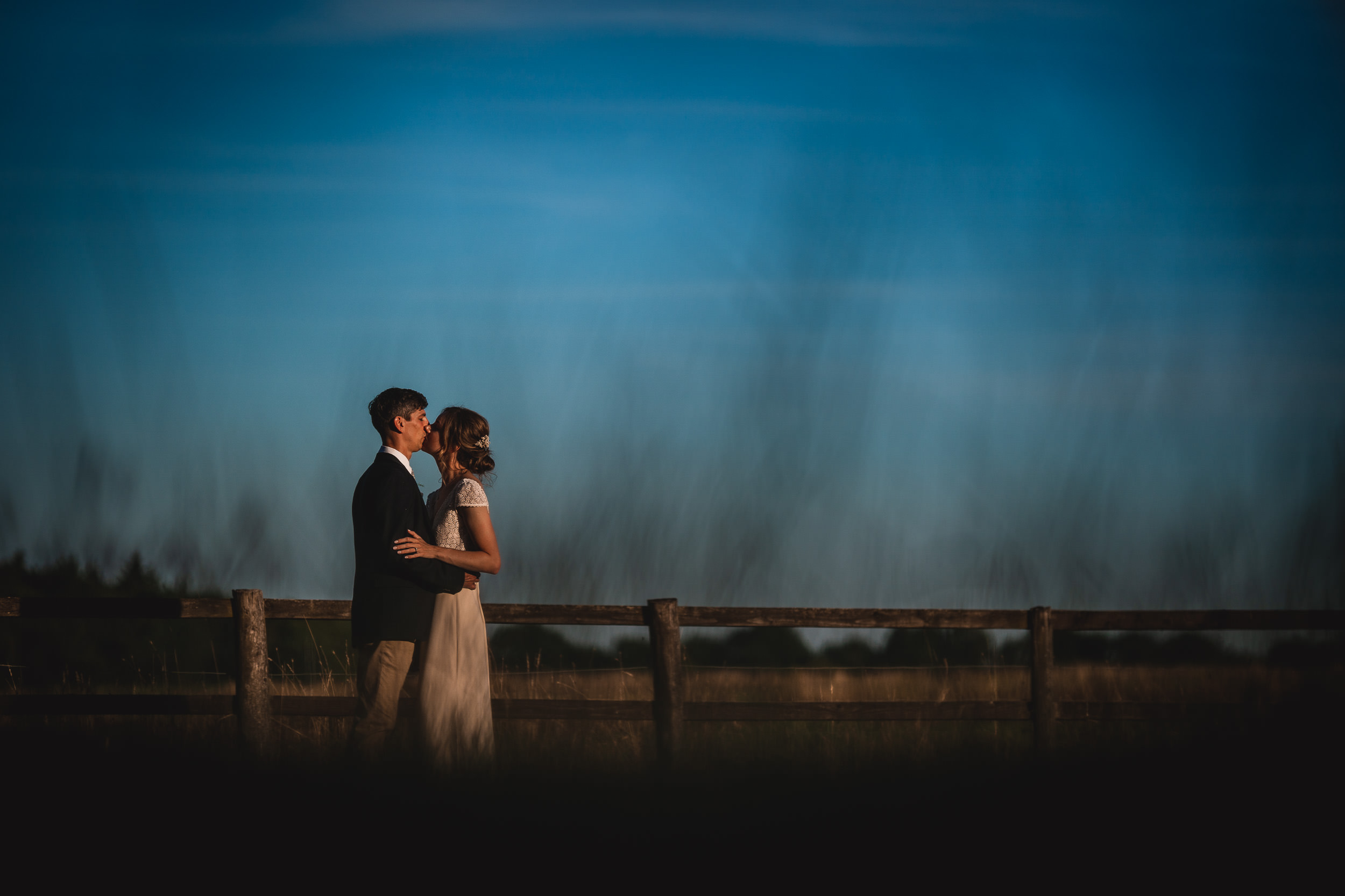 A bride and groom posing cheerfully for their wedding photographer in front of a fence at dusk.