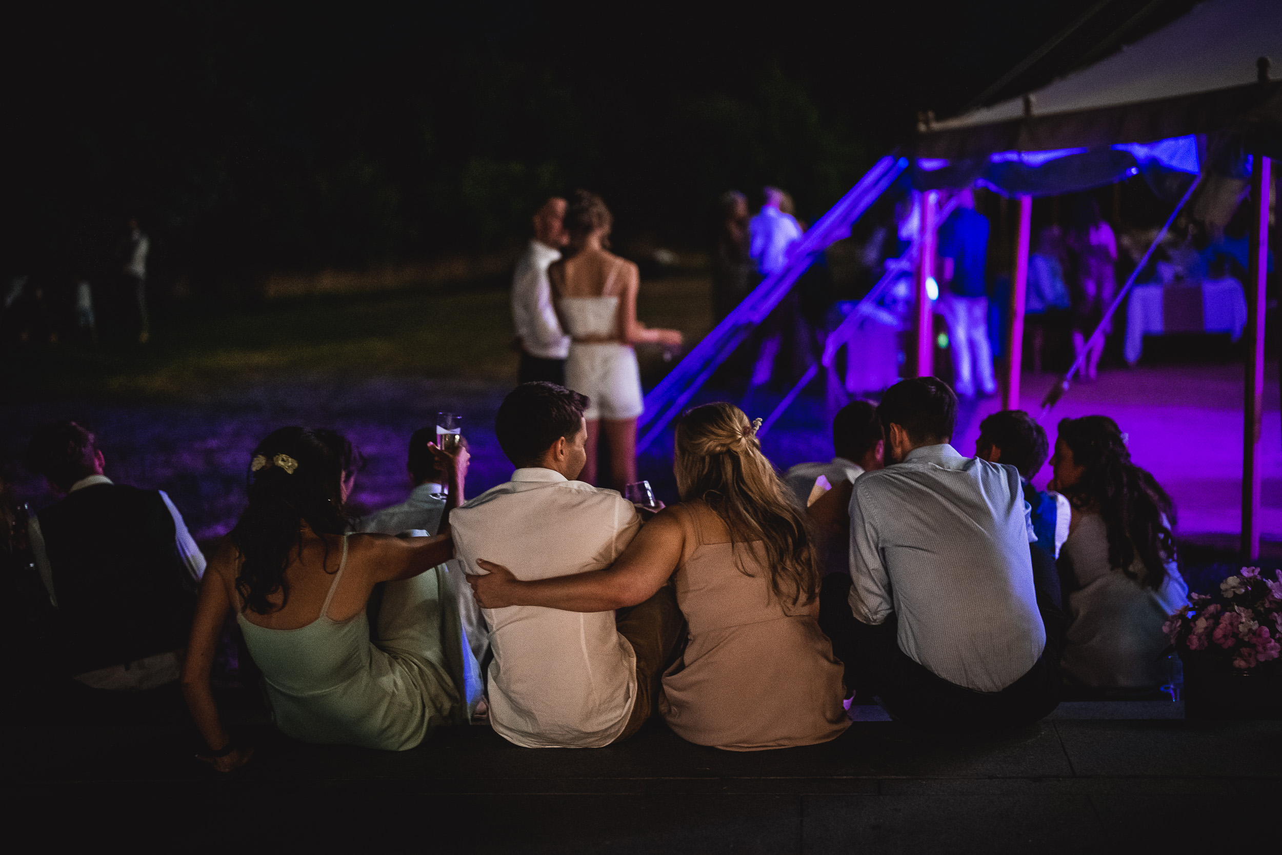 A group of people, including the groom and bride, sitting in front of a tent at night.