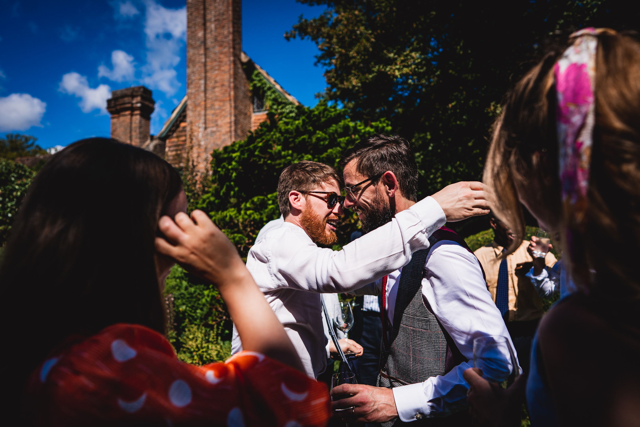 At a Surrey Wedding, a man is meticulously attaching a beard onto another man dressed in a suit at Ridge Farm.