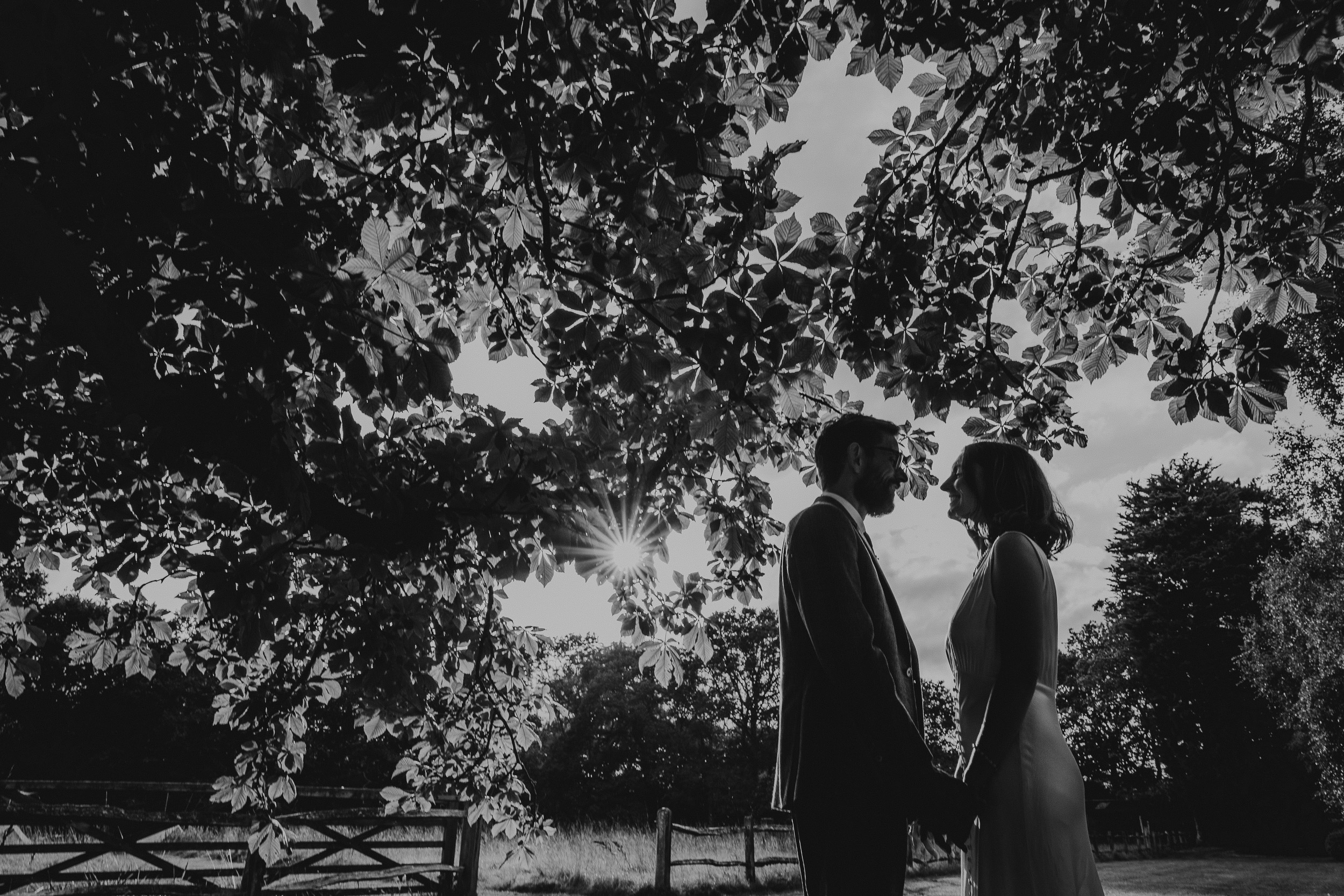 A black and white photo of a bride and groom standing under a tree during their Surrey wedding at Ridge Farm.