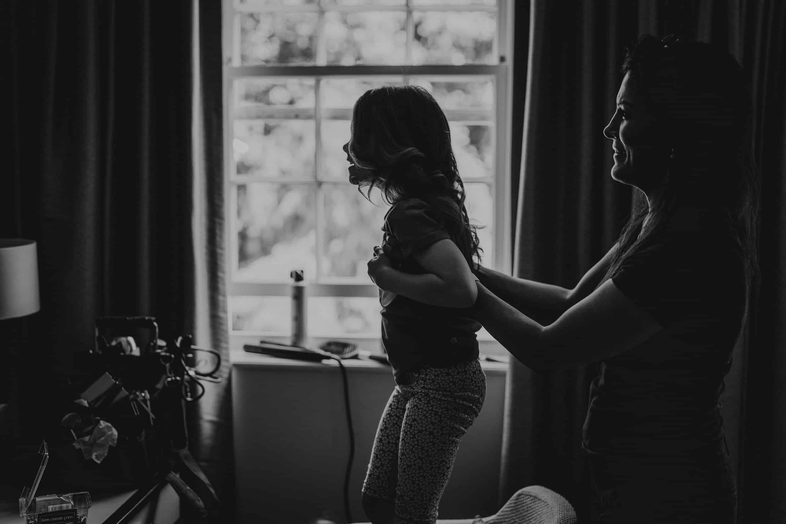 A woman helps a child stand on a chair in a dimly lit room with a window in the background.