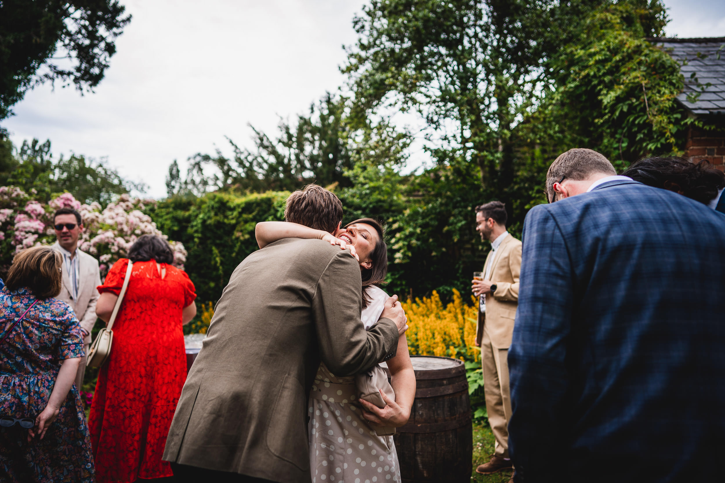 People hugging and socializing in a garden setting, surrounded by greenery and flowers.