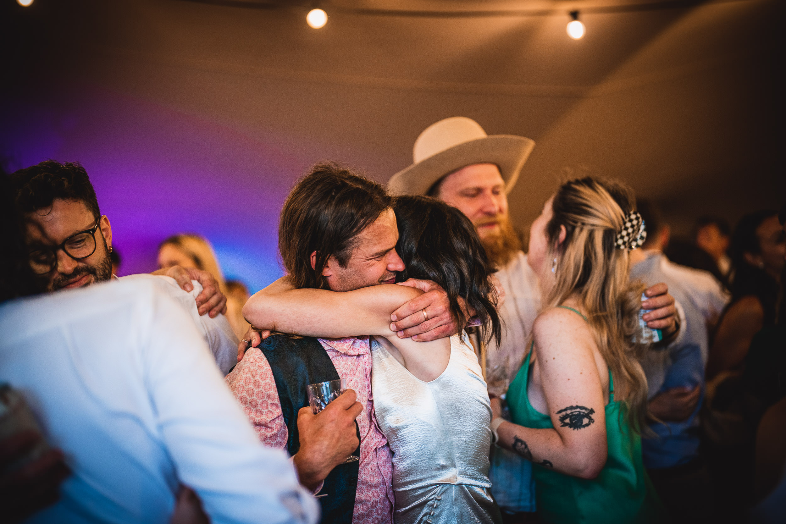 A group of people at a lively party. A couple in the center embraces; others stand nearby, one wearing a cowboy hat. The atmosphere is joyful with colorful lighting.