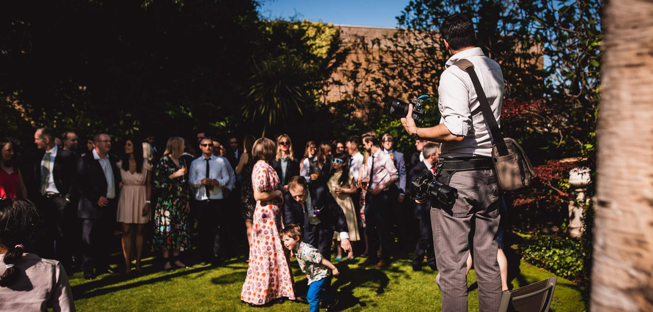 A photographer stands in the foreground taking photos of a group of adults and children gathered outdoors at a garden event on a sunny day.